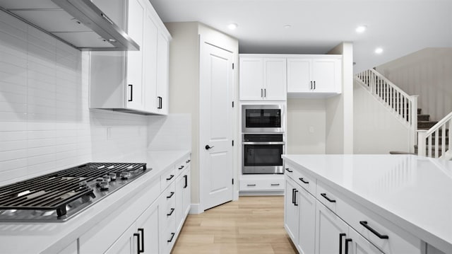 kitchen with appliances with stainless steel finishes, light wood-type flooring, tasteful backsplash, wall chimney exhaust hood, and white cabinetry