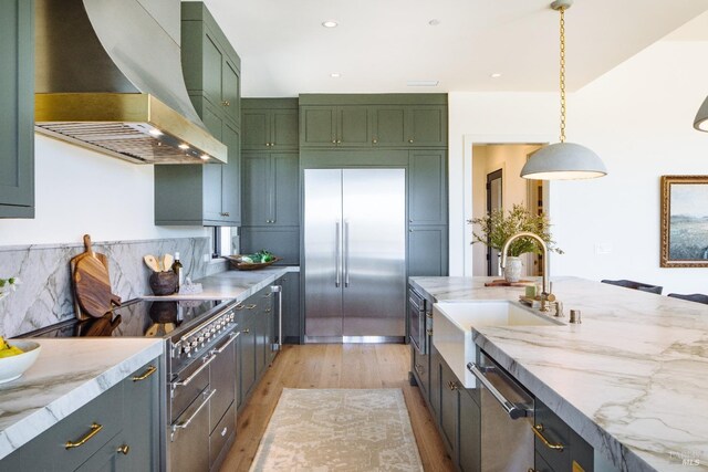 kitchen featuring wall chimney exhaust hood, green cabinetry, high end appliances, light hardwood / wood-style flooring, and decorative light fixtures