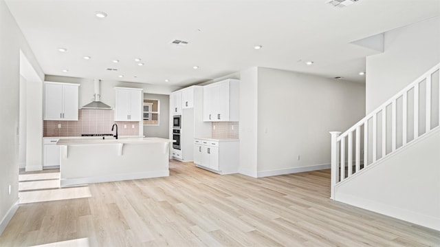 kitchen featuring wall chimney exhaust hood, light hardwood / wood-style flooring, an island with sink, decorative backsplash, and white cabinets