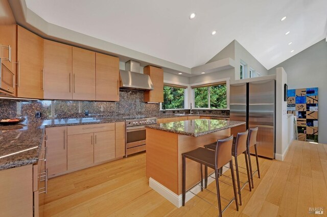 kitchen featuring appliances with stainless steel finishes, light hardwood / wood-style floors, dark stone counters, and wall chimney range hood