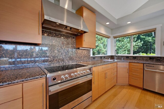 kitchen featuring light brown cabinets, light hardwood / wood-style flooring, wall chimney range hood, appliances with stainless steel finishes, and dark stone counters