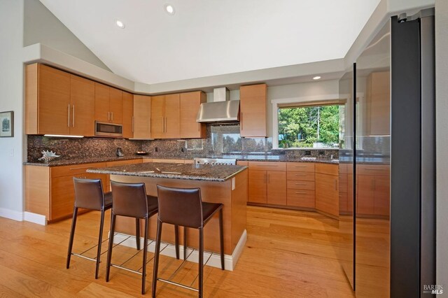 kitchen featuring a center island, vaulted ceiling, wall chimney exhaust hood, stainless steel appliances, and decorative backsplash