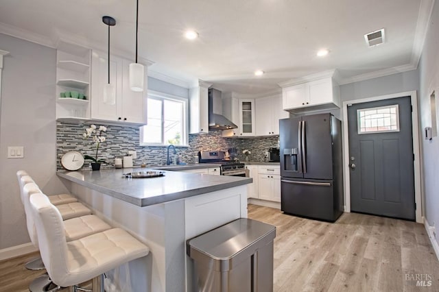 kitchen with appliances with stainless steel finishes, hanging light fixtures, white cabinets, kitchen peninsula, and wall chimney range hood