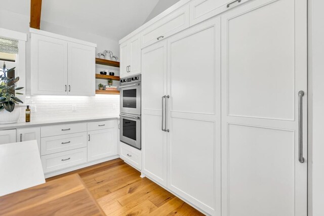 kitchen with backsplash, stainless steel double oven, white cabinets, and light wood-type flooring