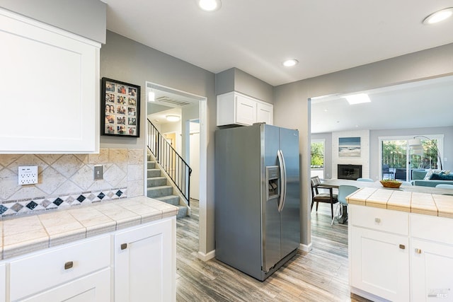 kitchen with tile countertops, stainless steel refrigerator with ice dispenser, a large fireplace, and light wood-type flooring