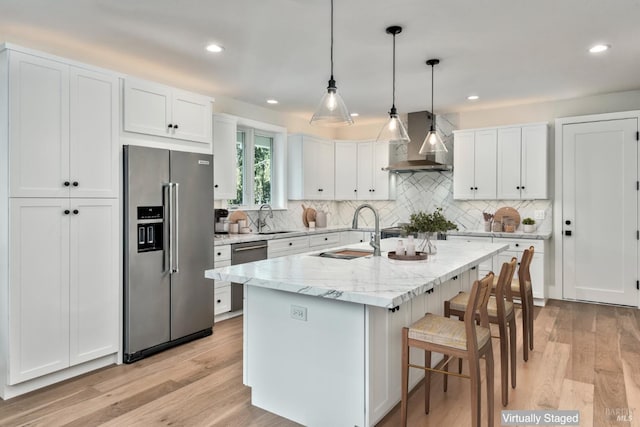 kitchen featuring wall chimney range hood, sink, white cabinetry, stainless steel appliances, and a kitchen island with sink