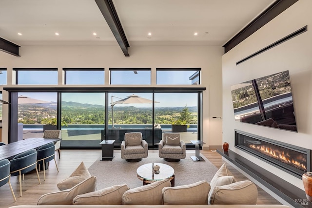 living area with recessed lighting, a towering ceiling, beamed ceiling, light wood finished floors, and a glass covered fireplace