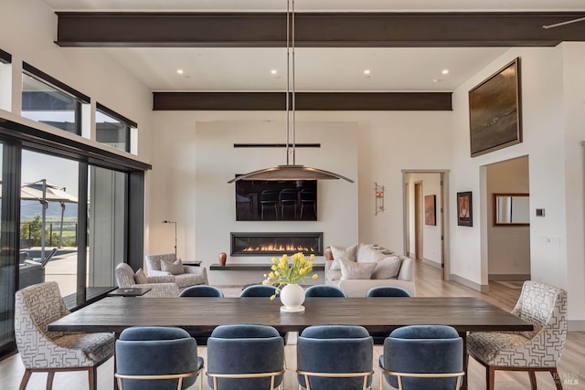 dining space with beam ceiling, a high ceiling, a glass covered fireplace, baseboards, and light wood-style floors