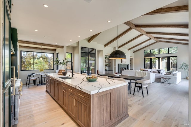 kitchen with light stone countertops, sink, vaulted ceiling with beams, light hardwood / wood-style floors, and an island with sink