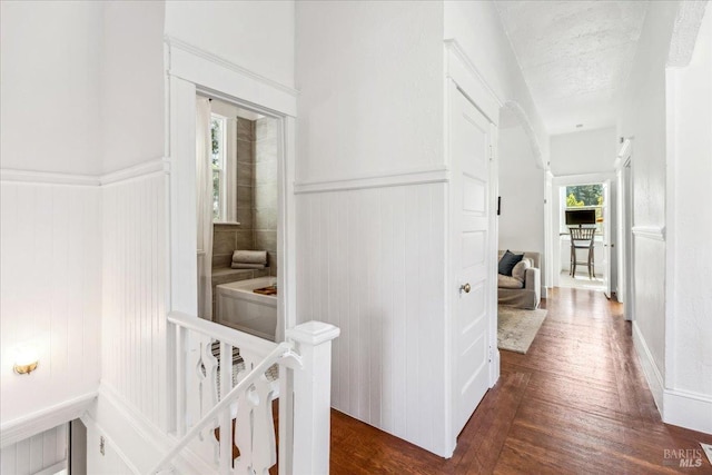 hallway with dark wood-style flooring, a wainscoted wall, a textured ceiling, and an upstairs landing