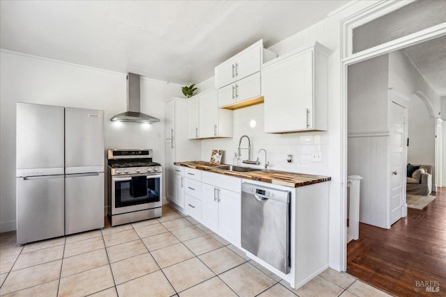 kitchen with light tile patterned floors, butcher block counters, appliances with stainless steel finishes, wall chimney range hood, and a sink