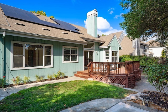 rear view of house featuring a deck, a yard, a shingled roof, solar panels, and a chimney