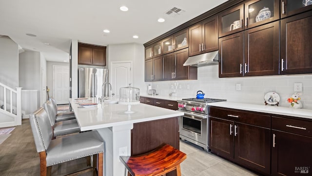 kitchen with dark brown cabinets, decorative backsplash, an island with sink, appliances with stainless steel finishes, and a kitchen bar