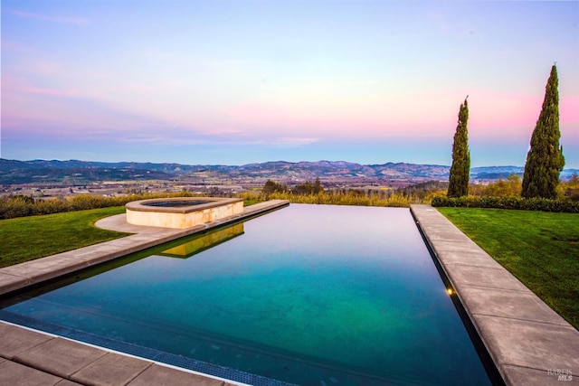 pool at dusk featuring a lawn, a mountain view, and an in ground hot tub