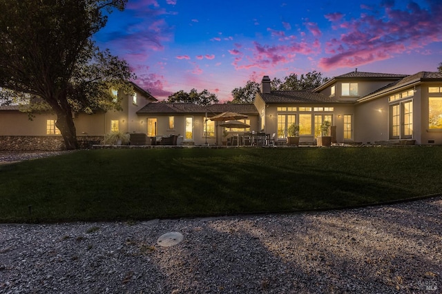 back of house at dusk featuring a lawn, a chimney, a patio, and stucco siding