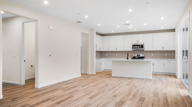 kitchen with an island with sink, stainless steel appliances, light hardwood / wood-style floors, decorative backsplash, and white cabinets