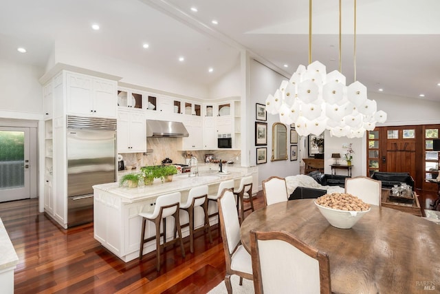 kitchen with light stone counters, hanging light fixtures, wall chimney range hood, built in fridge, and dark hardwood / wood-style floors