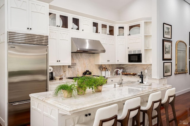 kitchen with wall chimney exhaust hood, dark wood-type flooring, sink, light stone countertops, and built in appliances