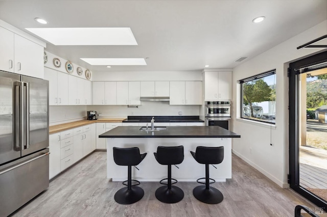 kitchen featuring a skylight, sink, a center island with sink, white cabinets, and appliances with stainless steel finishes