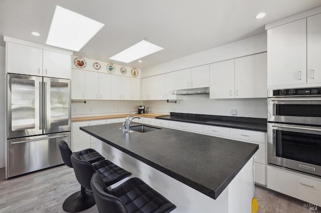 kitchen featuring a skylight, stainless steel appliances, a kitchen island with sink, sink, and white cabinets