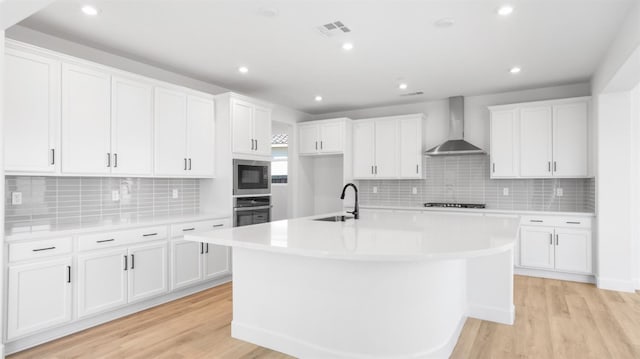 kitchen featuring wall chimney range hood, light wood-type flooring, stainless steel appliances, white cabinetry, and a kitchen island with sink