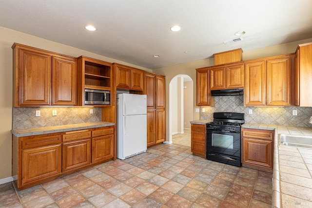 kitchen featuring tasteful backsplash, tile countertops, stainless steel microwave, black gas stove, and white refrigerator