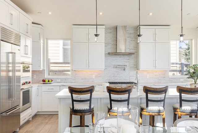 kitchen with wall chimney range hood, stainless steel appliances, decorative light fixtures, white cabinets, and a breakfast bar