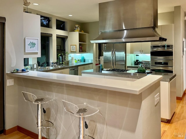 kitchen with kitchen peninsula, island exhaust hood, white cabinets, appliances with stainless steel finishes, and light hardwood / wood-style floors