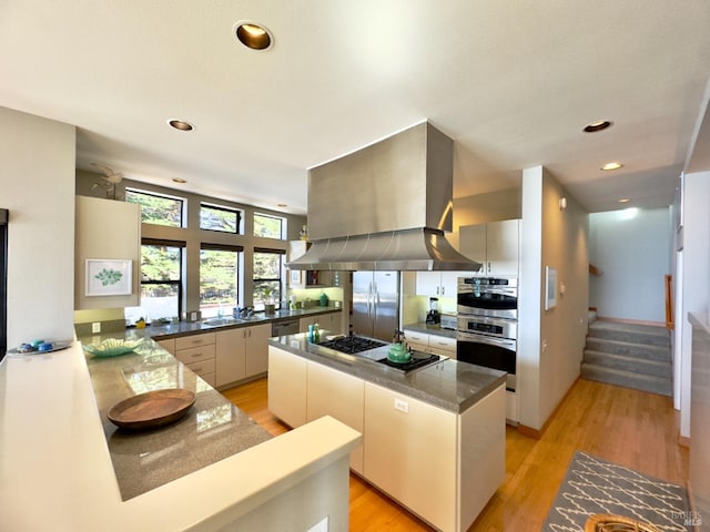 kitchen with kitchen peninsula, appliances with stainless steel finishes, island range hood, light wood-type flooring, and sink