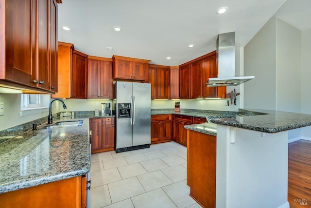 kitchen featuring sink, wall chimney range hood, kitchen peninsula, stainless steel fridge, and dark stone counters