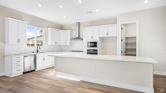 kitchen featuring wall chimney range hood, a center island, stainless steel appliances, white cabinets, and light hardwood / wood-style flooring