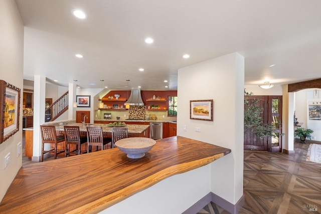 kitchen featuring dishwasher, wall chimney range hood, kitchen peninsula, dark parquet flooring, and a breakfast bar