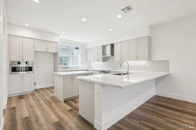 kitchen with oven, a peninsula, a sink, visible vents, and wall chimney exhaust hood