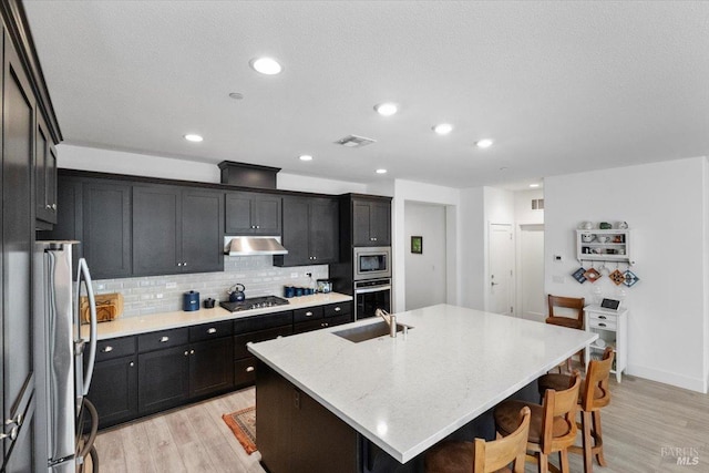 kitchen with light stone countertops, light wood-type flooring, an island with sink, tasteful backsplash, and stainless steel appliances