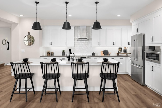 kitchen featuring dark hardwood / wood-style flooring, wall chimney range hood, a kitchen island with sink, and appliances with stainless steel finishes