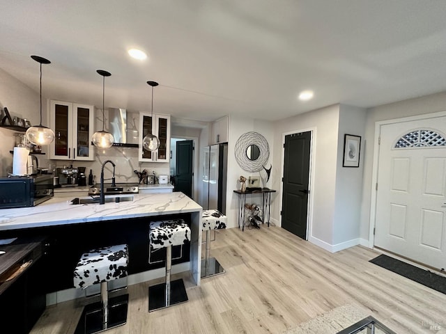 kitchen with a sink, freestanding refrigerator, wall chimney exhaust hood, light wood finished floors, and glass insert cabinets