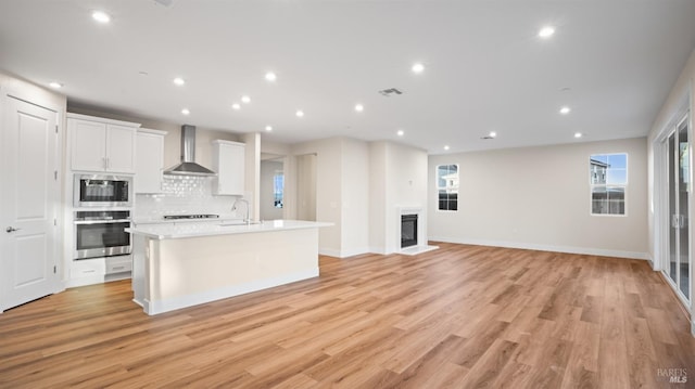 kitchen featuring wall chimney range hood, white cabinetry, an island with sink, built in microwave, and oven