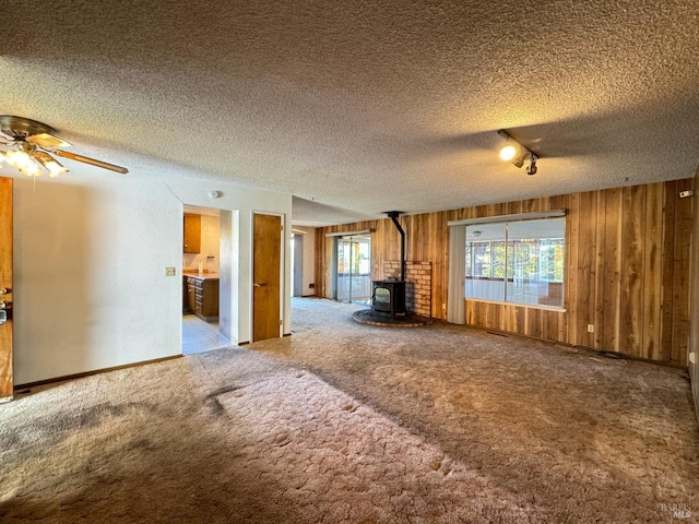 unfurnished living room with wood walls, carpet floors, a wood stove, ceiling fan, and a textured ceiling