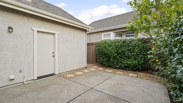 view of exterior entry featuring roof with shingles, a patio area, fence, and stucco siding