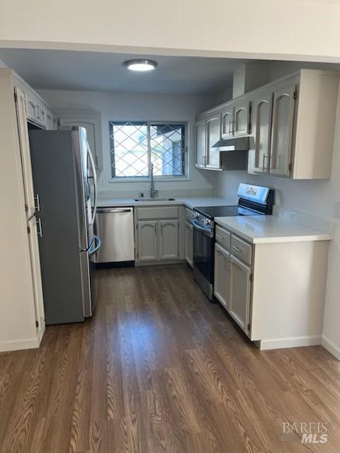 kitchen featuring stainless steel appliances, under cabinet range hood, light countertops, dark wood-style flooring, and a sink