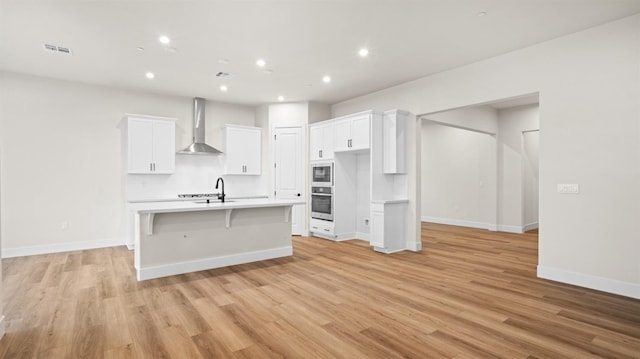 kitchen with white cabinets, an island with sink, wall chimney range hood, and oven