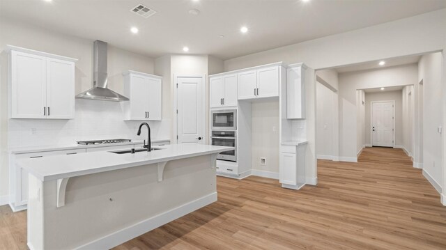 kitchen with white cabinetry, sink, stainless steel oven, a center island with sink, and wall chimney exhaust hood