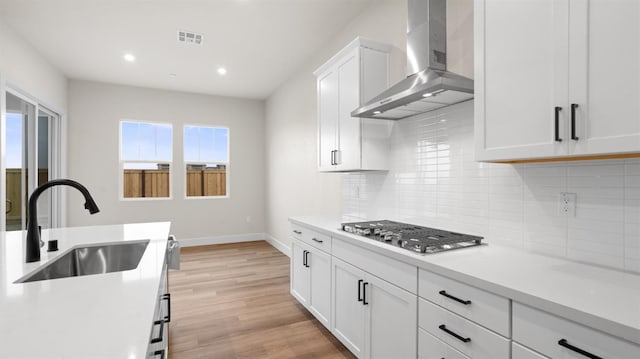 kitchen featuring wall chimney range hood, sink, light hardwood / wood-style flooring, white cabinetry, and stainless steel gas cooktop