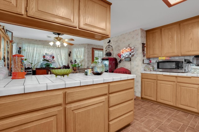 kitchen featuring wallpapered walls, tile countertops, ceiling fan, stainless steel microwave, and brick patterned floor