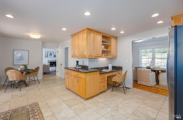 kitchen with stainless steel fridge, dark stone counters, light brown cabinets, light tile patterned floors, and a tiled fireplace