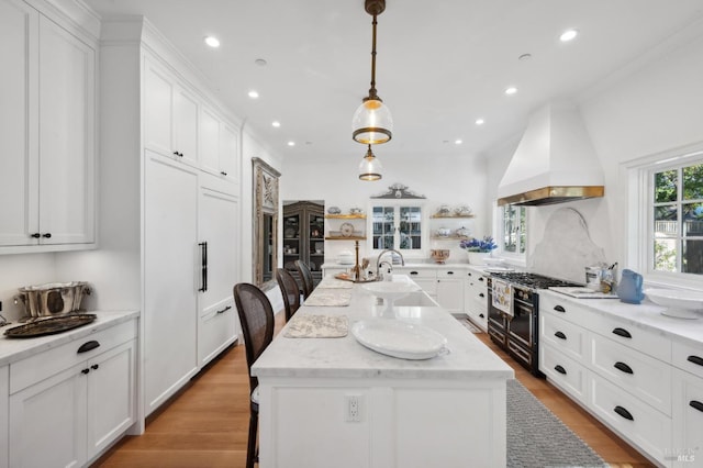 kitchen featuring custom exhaust hood, a breakfast bar, an island with sink, stainless steel range, and light stone counters