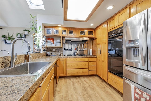 kitchen with dobule oven black, a skylight, under cabinet range hood, stainless steel refrigerator with ice dispenser, and a sink