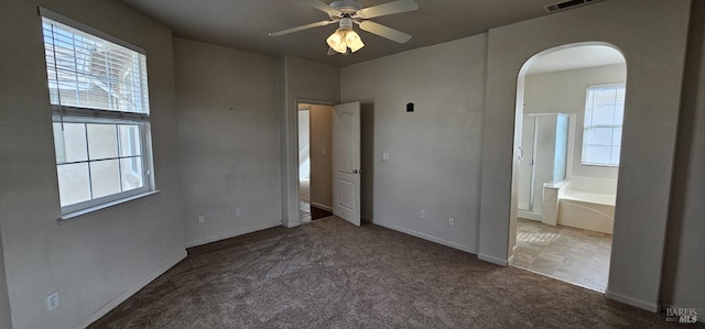 unfurnished bedroom featuring ceiling fan, dark carpet, ensuite bathroom, and multiple windows