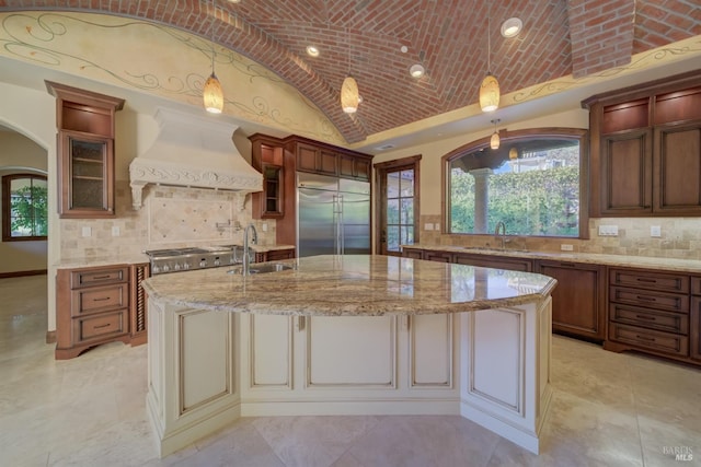 kitchen featuring custom exhaust hood, stainless steel appliances, decorative light fixtures, lofted ceiling, and an island with sink