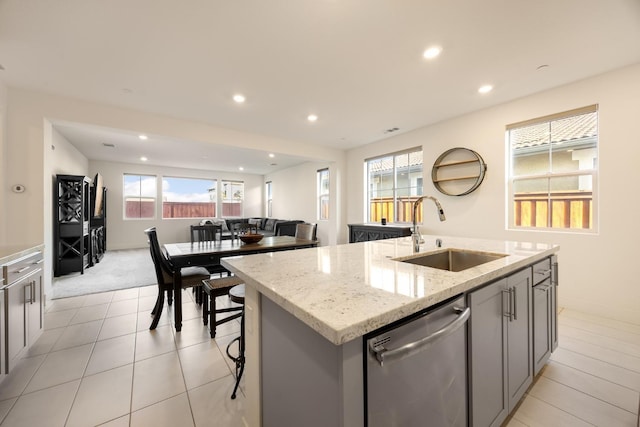 kitchen with a sink, dishwasher, gray cabinetry, a center island with sink, and light stone countertops